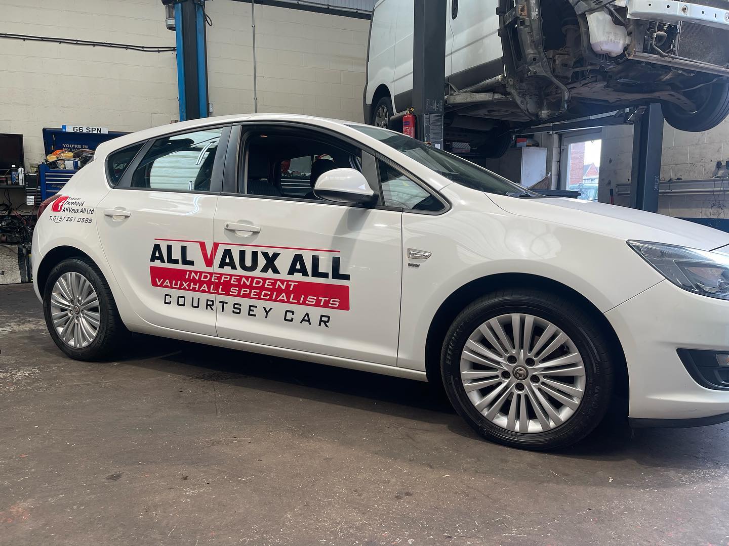 White Vauxhall Astra courtesy car in All Vaux All livery inside the workshop, with a van raised on a lift behind it